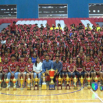 Large group of people in matching jerseys seated in bleachers with numerous trophies displayed in front.