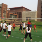 People playing volleyball on a green court surrounded by modern brick buildings and grassy areas.