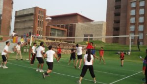 People playing volleyball on a green court surrounded by modern brick buildings and grassy areas.
