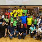 Group of people, including children in colorful sportswear with medals, posing indoors on a wooden floor.