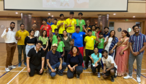 Group of people, including children in colorful sportswear with medals, posing indoors on a wooden floor.