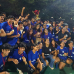 Group of students in blue shirts celebrating with medals and a trophy outdoors at night.