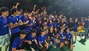 Group of students in blue shirts celebrating with medals and a trophy outdoors at night.