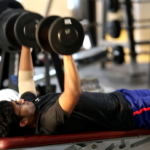 Person lying on a bench in a gym, lifting dumbbells with both arms, wearing glasses and workout clothes.