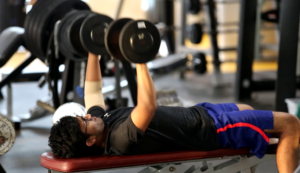 Person lying on a bench in a gym, lifting dumbbells with both arms, wearing glasses and workout clothes.