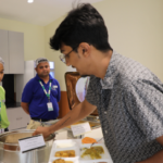 Man serving food from a buffet counter while two staff members in uniforms assist in the background.