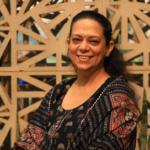 Smiling woman in a patterned dress sitting in front of a geometric lattice background.