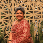 Smiling woman in a red saree with a patterned background and green plants in the foreground.