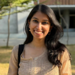 Smiling woman with long dark hair wearing a beige top, standing outdoors in sunlight near trees.