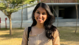 Smiling woman with long dark hair wearing a beige top, standing outdoors in sunlight near trees.