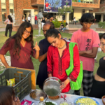 Group of people preparing and enjoying food at an outdoor event on a sunny day with a campus backdrop.