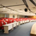 Lecture hall with rows of red chairs, white desks, a wooden stage, and ceiling-mounted lights.