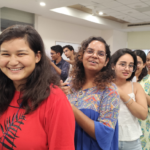 A group of smiling people standing in a line indoors, some holding pens and papers.