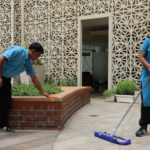 Two workers in blue uniforms cleaning an outdoor area with a mop and cloth near a patterned wall.