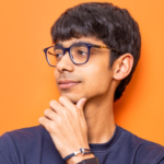 Young man with glasses and smartwatch, wearing a navy shirt, posing against an orange background.