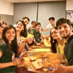 A group of smiling people crafting yellow paper decorations around a table in a well-lit room.