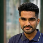 Smiling man with short black hair wearing a navy polo shirt, blurred windowed background.