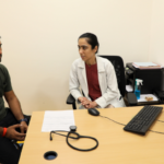 Doctor and patient seated at a desk with a stethoscope, computer, and documents in a clinical office.