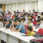 Students seated in a lecture hall, listening attentively with notebooks and laptops on desks.