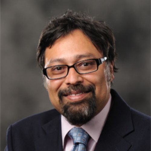 Smiling man with glasses, beard, and mustache wearing a suit, tie, and pink shirt on a dark background.