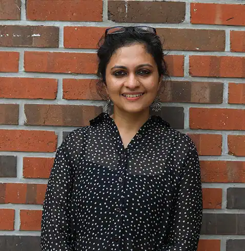 Smiling woman with dark curly hair, wearing a black polka dot shirt, standing against a brick wall.