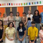 Group of people posing indoors, colorful flags hanging above, orange and gray panels in the background.