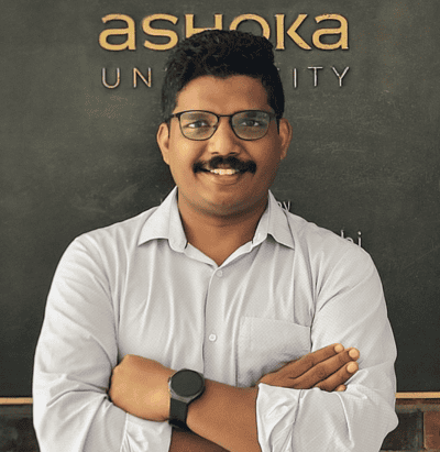 Smiling man with glasses and folded arms, standing in front of an Ashoka University sign.