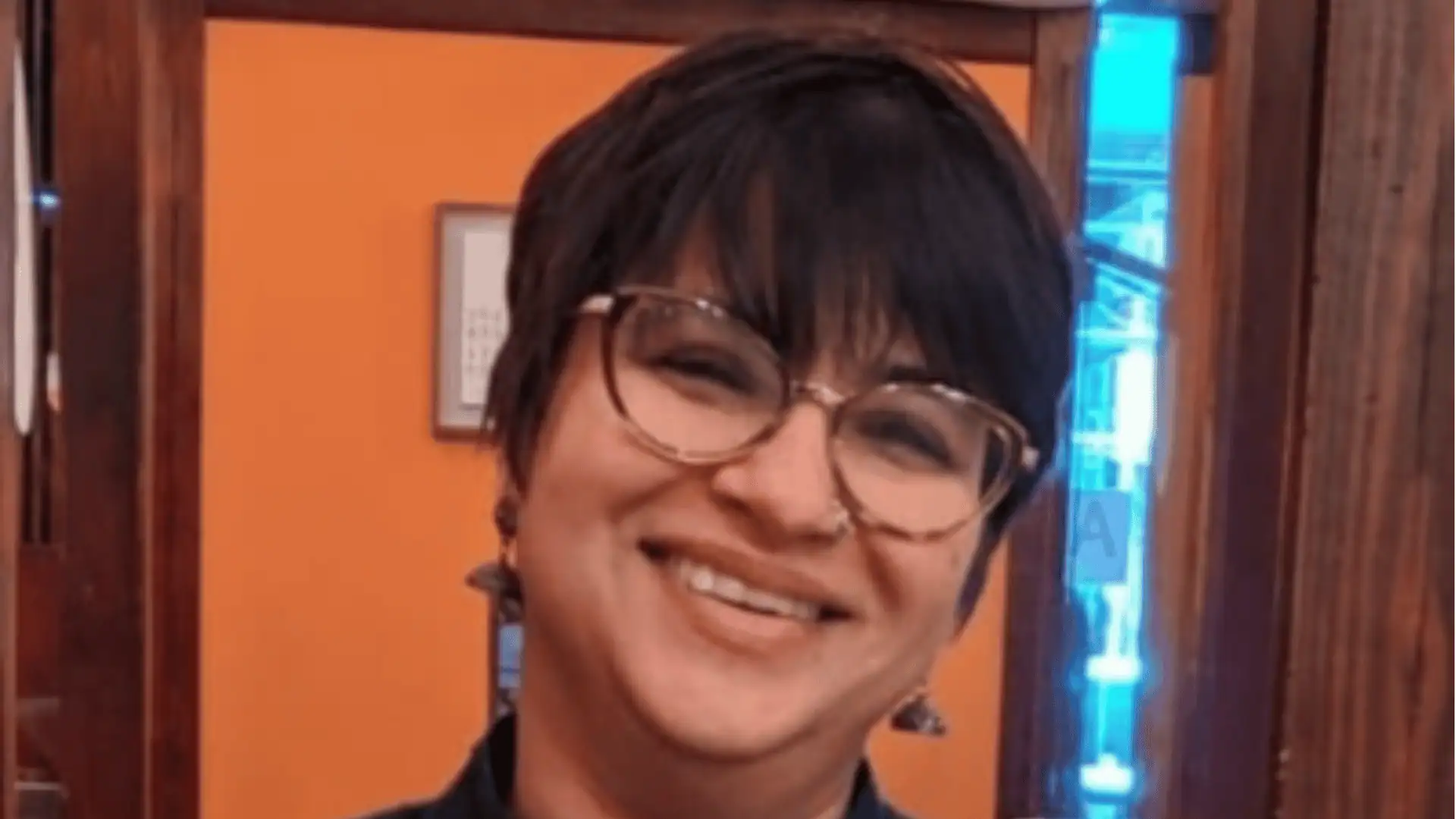 Smiling person with short dark hair, glasses, nose ring, and earrings, in front of an orange wall.