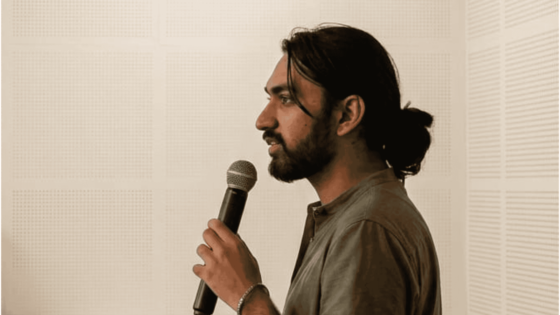 Person with a microphone, wearing a green shirt, speaking in front of a light perforated wall.