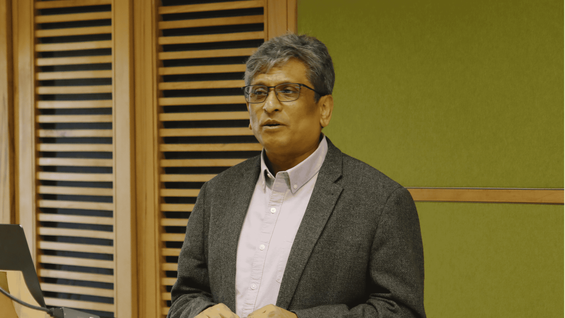 Man with glasses and gray hair speaking, wearing a blazer and shirt, wooden panels in the background.
