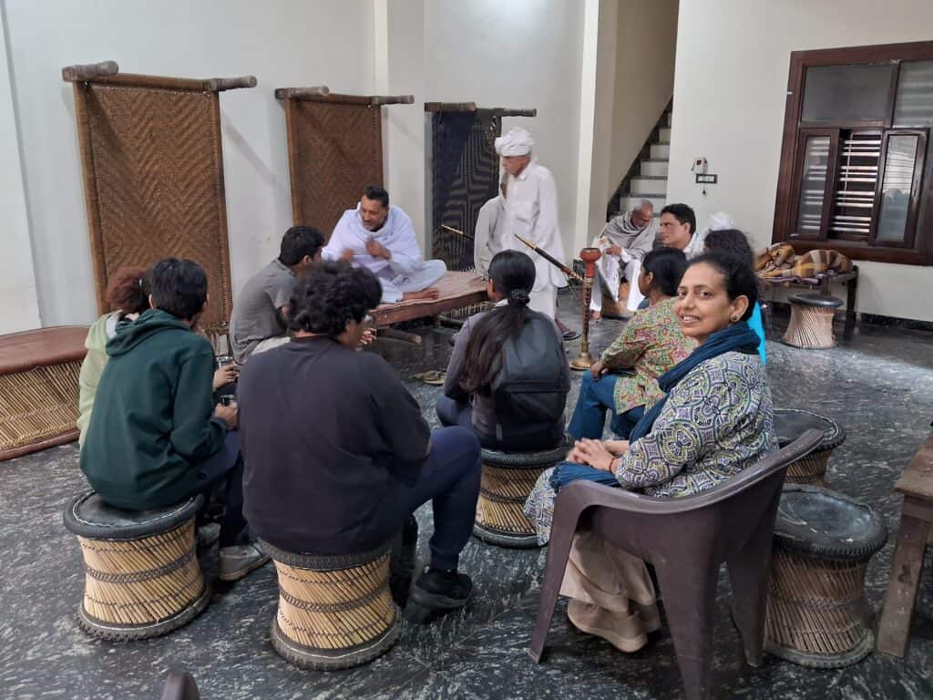 Group seated indoors on woven stools, two men in traditional attire talking on a charpai in background.