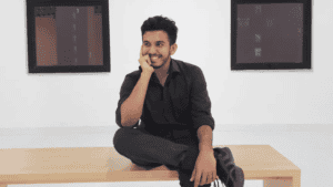 Man sitting cross-legged on a wooden bench, smiling, wearing a black outfit, with two windows behind.