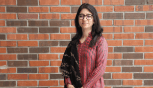 Woman with glasses, wearing a red patterned kurta and black shawl, standing against a brick wall.