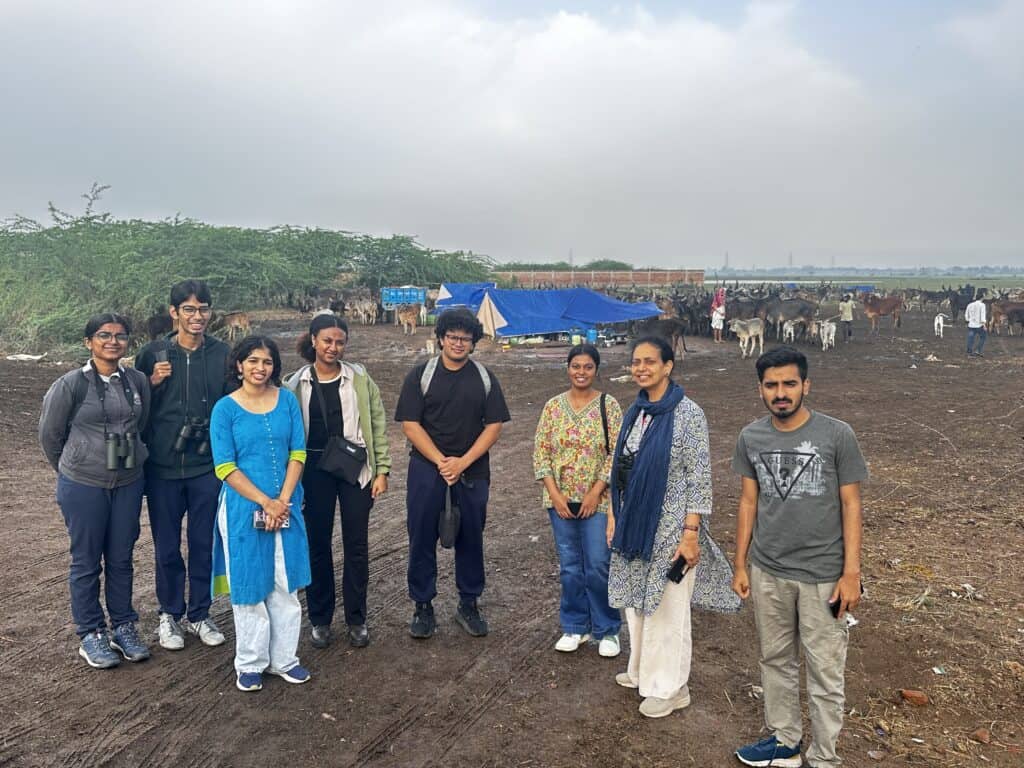 Group of eight people standing outdoors on a dirt field with cattle, trees, and tents in the background.