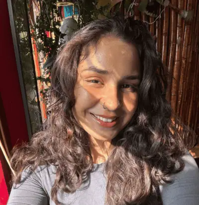Smiling person with long curly hair and a nose ring, sunlight casting shadows on their face.