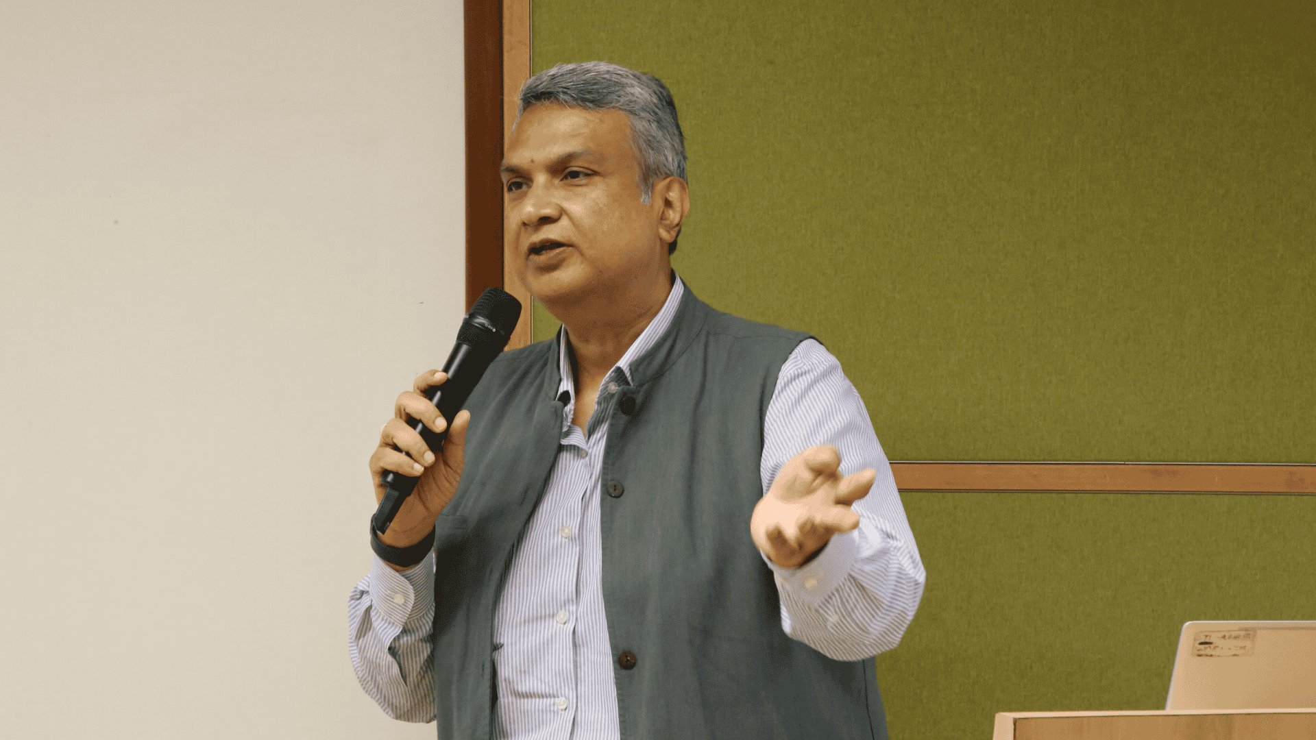 Man speaking into a microphone, gesturing with one hand, standing in front of a green background.