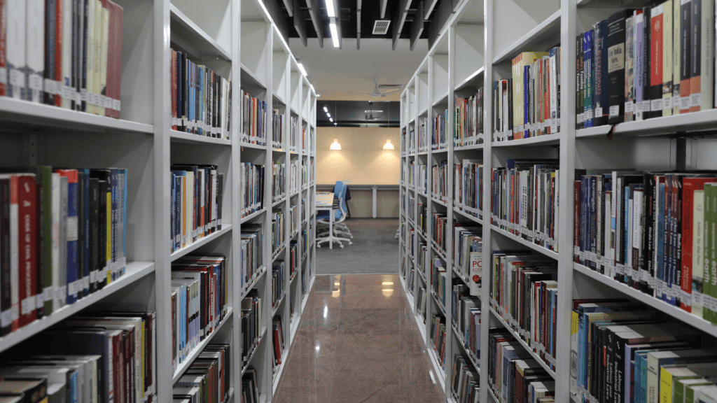 Library aisle with white shelves filled with books, leading to a study area with tables and chairs.
