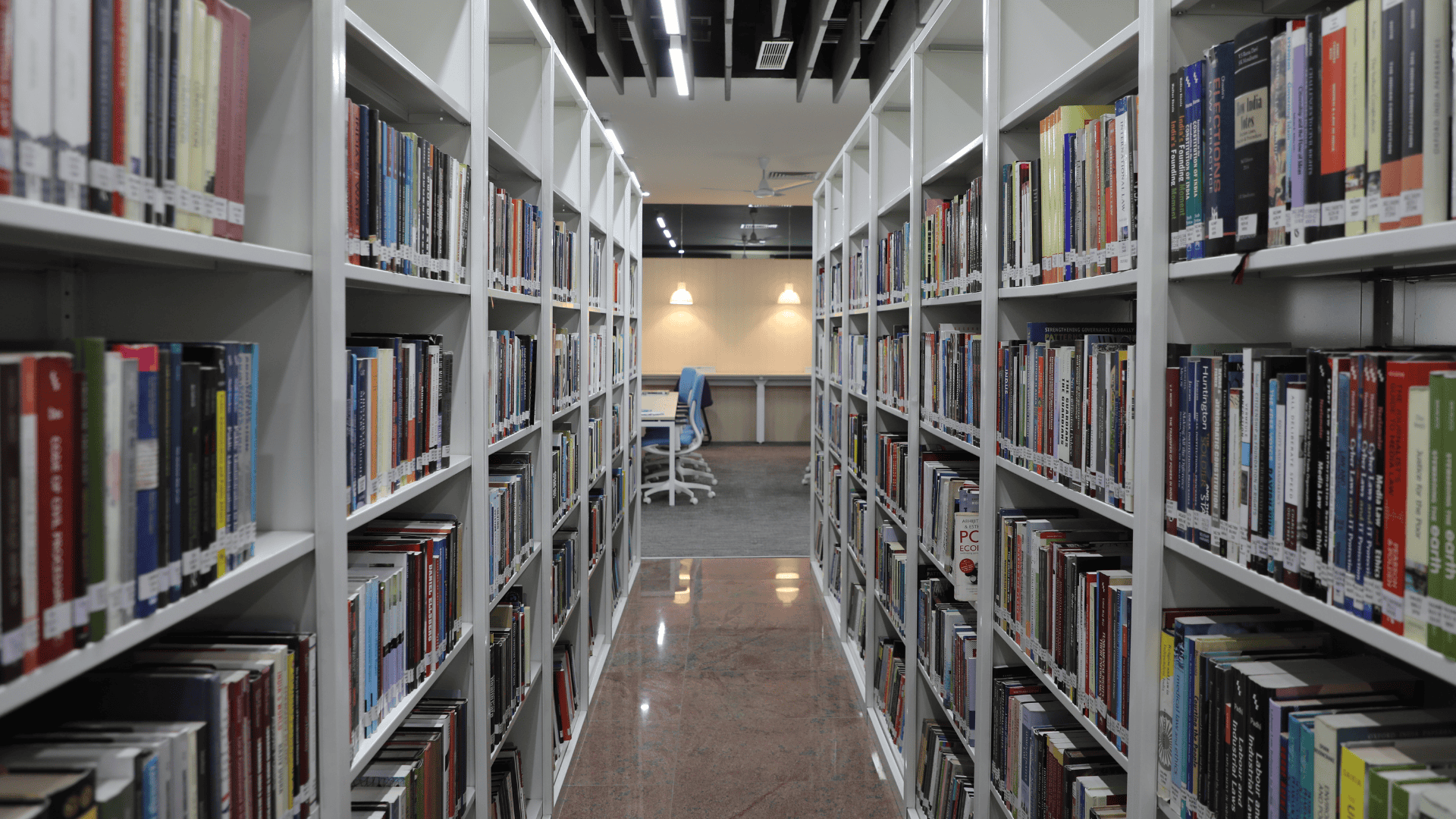 Library aisle with white shelves filled with books, leading to a study area with tables and chairs.