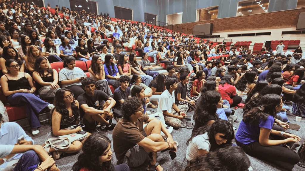 Crowded auditorium with seated students attentively facing forward, some on chairs, others on the floor.