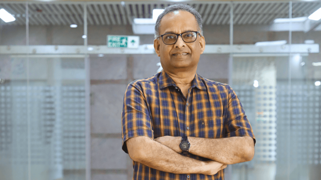 Man in a checkered shirt and glasses standing with arms crossed in a modern glass-walled space.