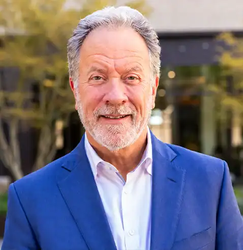 Smiling man with gray hair and beard wearing a blue suit and white shirt outdoors.
