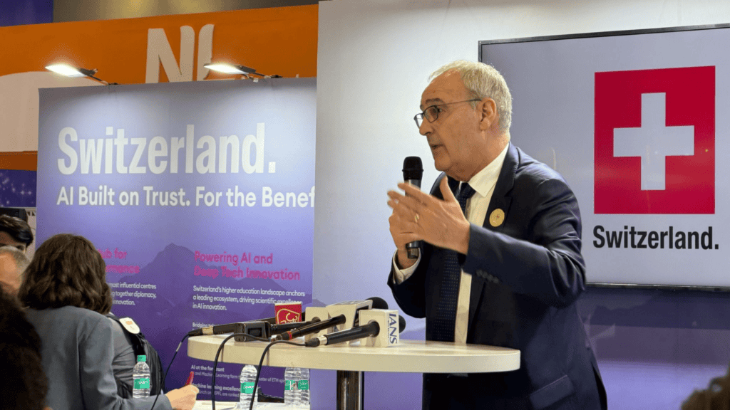 Man in a suit speaking into a microphone at a podium, Switzerland banner and logo in the background.