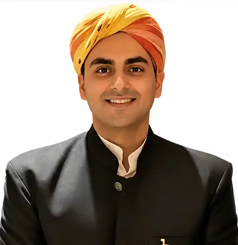 Smiling man wearing a colorful turban and a black traditional outfit against a white background.