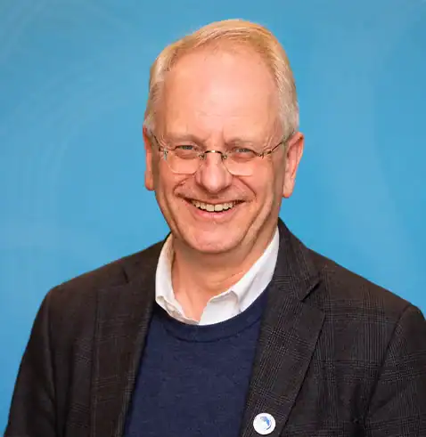 Smiling man with glasses, wearing a dark blazer, blue sweater, and white shirt against a blue background.