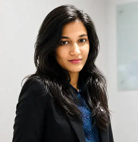 Woman with long dark hair wearing a black blazer and blue shirt, standing against a light background.