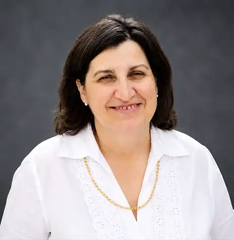 Smiling woman with shoulder-length dark hair, white blouse, and gold necklace, gray background.