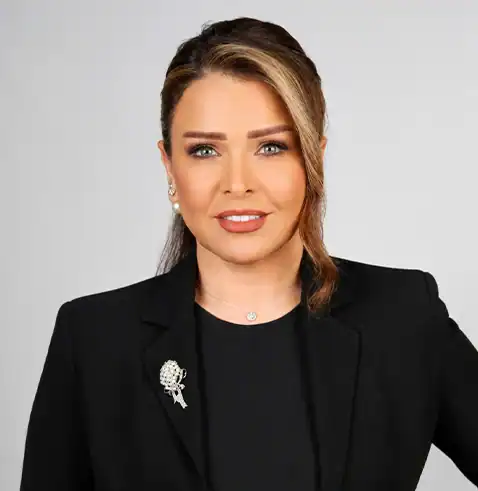 Woman with styled hair wearing a black blazer, pearl earrings, and a brooch, smiling confidently.