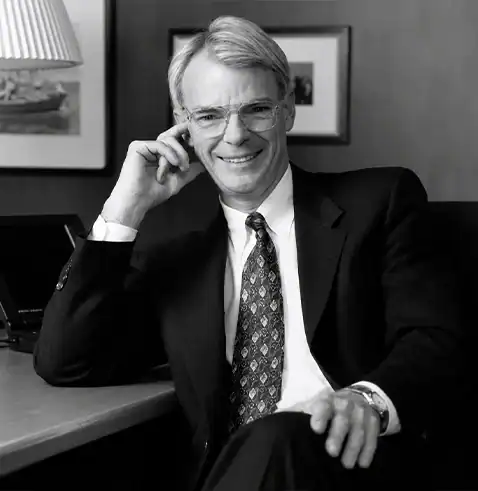 Smiling man in a suit and tie, seated at a desk, with framed art and a lamp in the background.