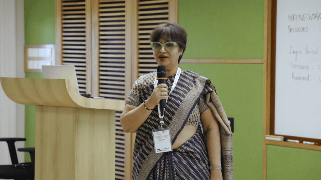 Professor Nandini speaking into a microphone near a wooden podium and whiteboard.