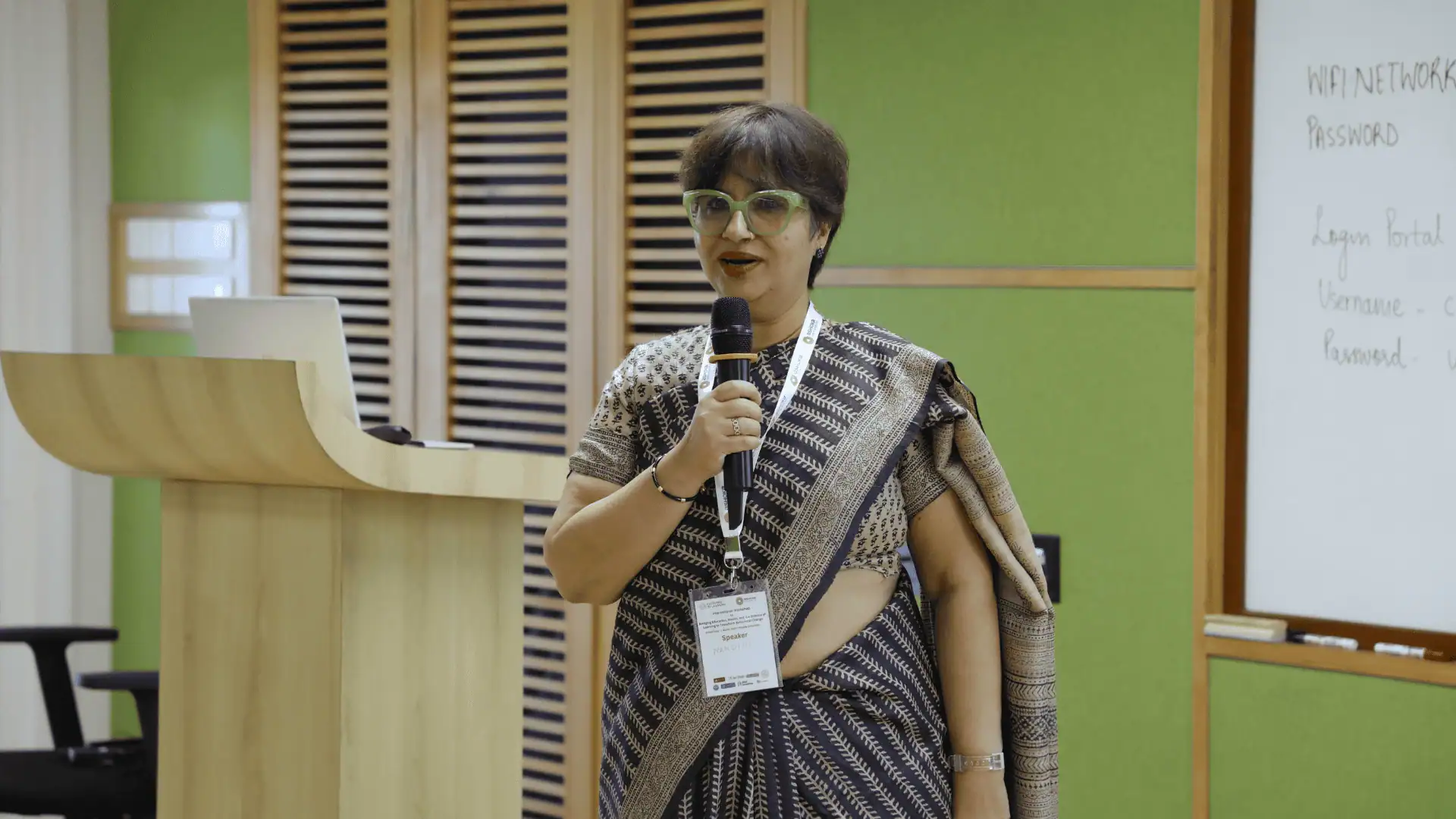 Professor Nandini speaking into a microphone near a wooden podium and whiteboard.